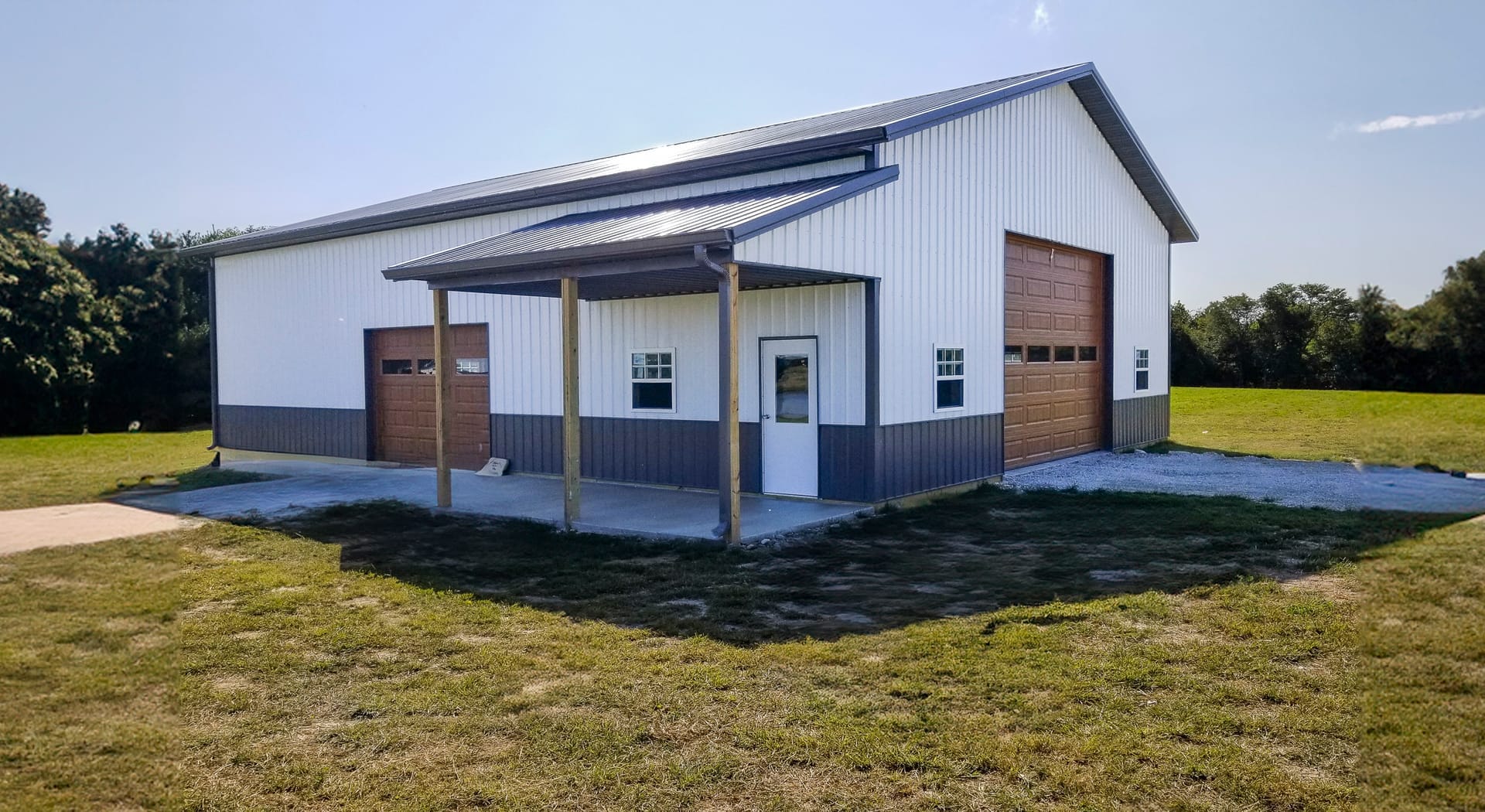 The image depicts a post-frame building from Built-Mor Buildings, featuring a gable roof and metal siding in white and gray. The structure has three large overhead doors, suggesting use as a garage or storage facility. A small covered entryway, supported