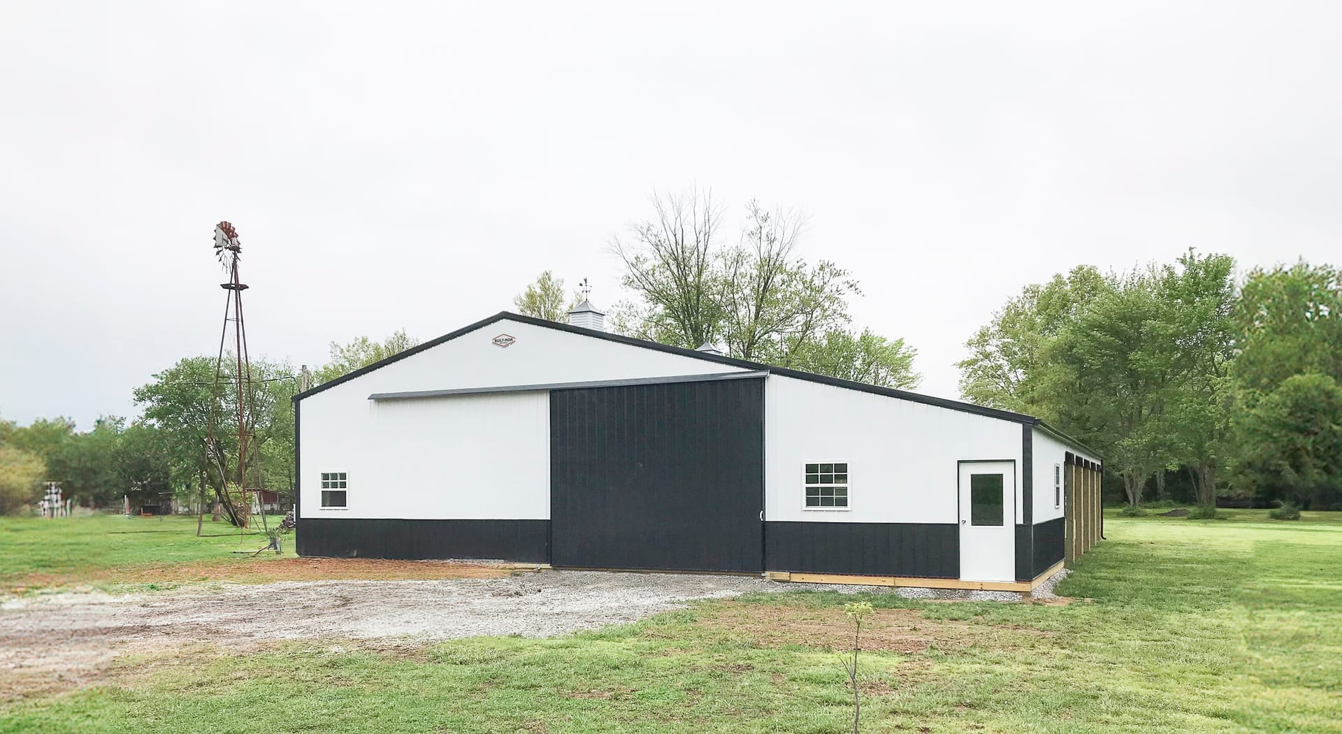 A post-frame construction building by Built-Mor Buildings is shown in a rural setting. The structure has a classic barn-like design with a gable roof and large sliding doors on the front facade. It features a two-tone color scheme with white upper walls a