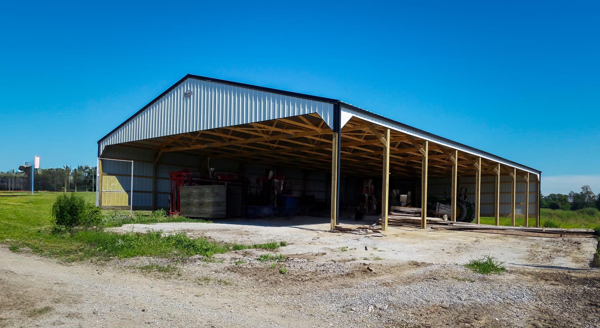 The image depicts a large post-frame construction building, likely a storage or agricultural facility, with an open side displaying a spacious interior. The structure features a metal roof and siding, supported by wooden posts and beams. The roof has a ga