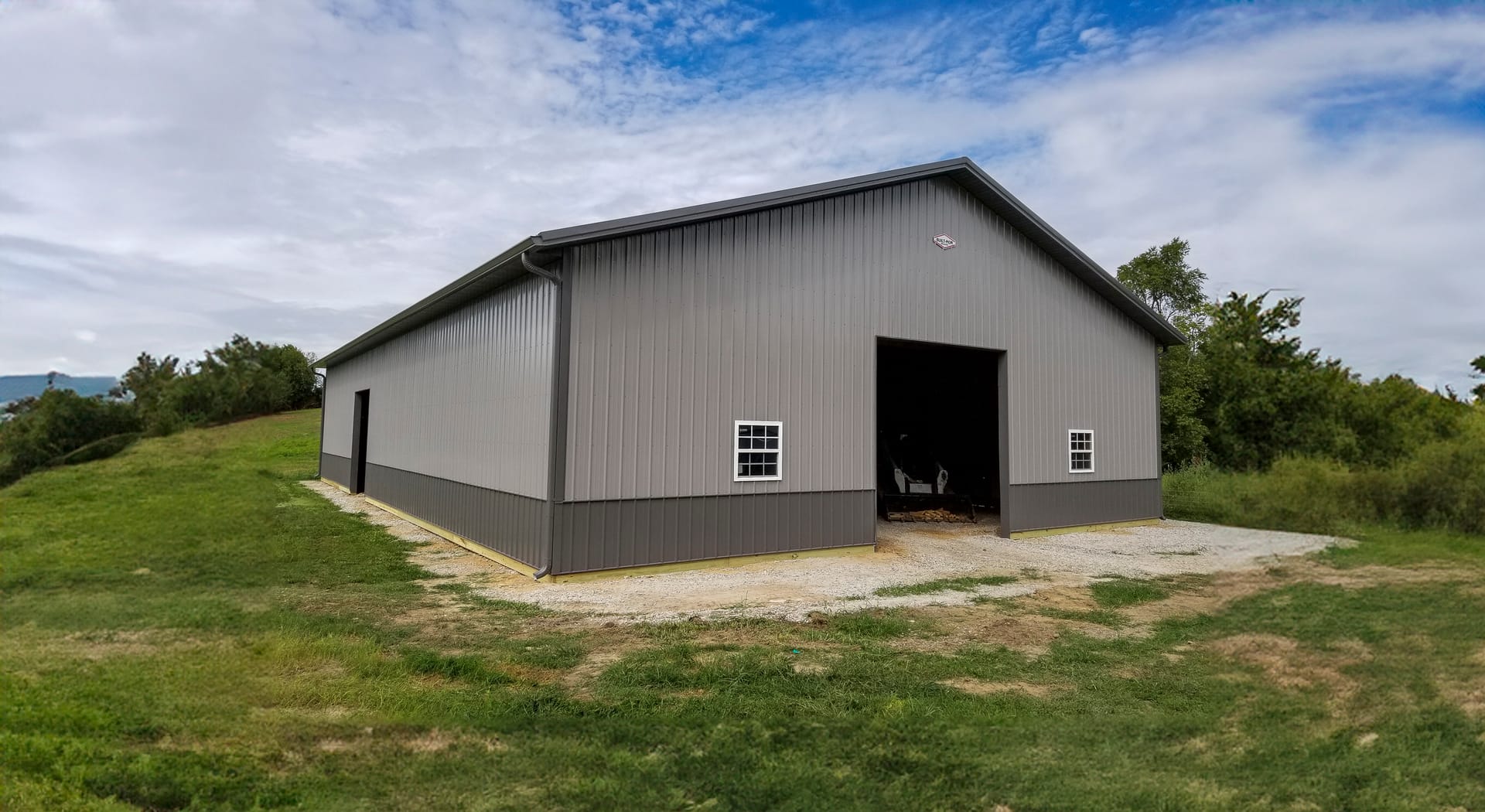 The image shows a post-frame construction building by Built-Mor Buildings, featuring a simple gable roof and metal siding. The exterior is divided into two tones, with darker panels at the bottom and lighter panels above. The structure includes two small