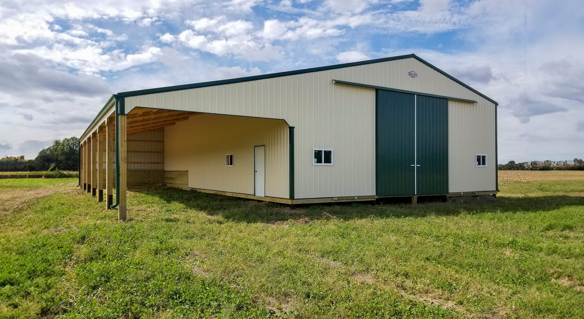 A large post-frame construction building with beige siding and a dark green roof, featuring an open-sided section supported by wooden posts. It includes a set of wide, double sliding doors on the front, and several small windows along the walls. The build
