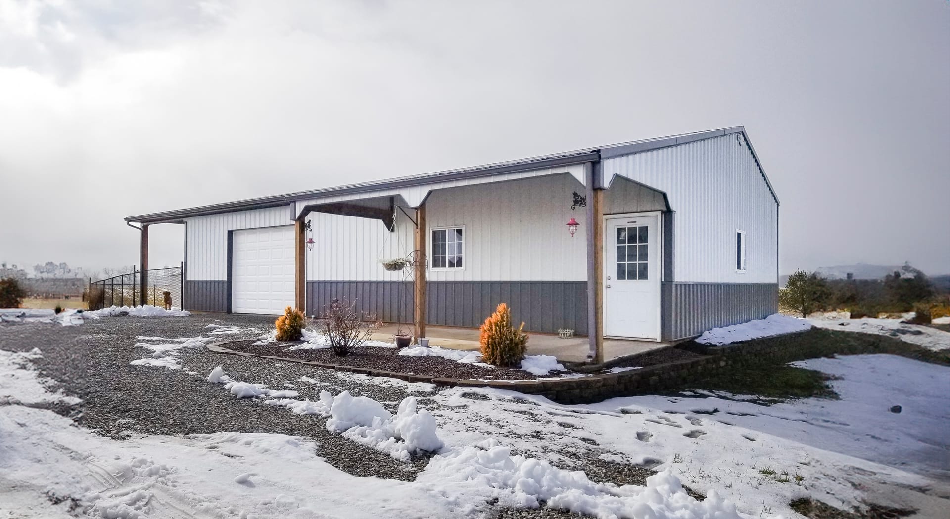A modern post-frame building set in a snowy landscape, likely used for storage or as a workshop. It features a gable roof and a two-tone exterior with vertical siding—grey on the lower half and white above. The structure includes a large garage door and a