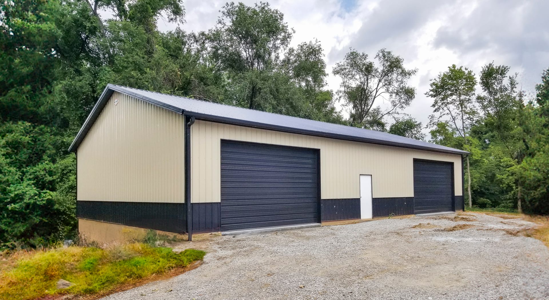 A post-frame construction building with beige metal siding and a gabled roof, set against a wooded backdrop. The structure features three large black overhead garage doors and one standard white entry door, indicating its use as a storage or workshop faci