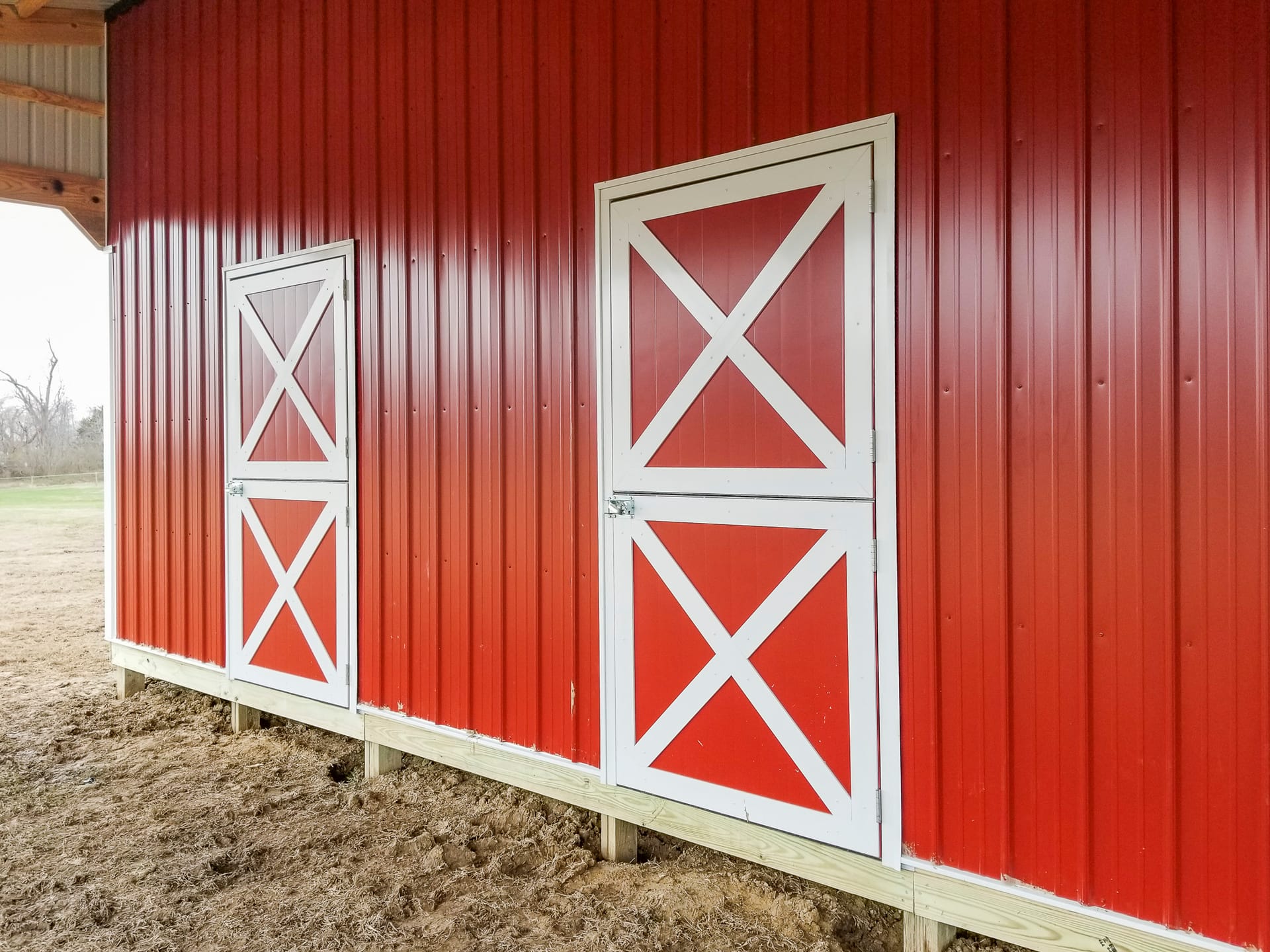 The image shows a post-frame building by Built-Mor Buildings, featuring red vertical metal siding with white diagonal cross-braced doors, typical of barn-style design. The exterior wall includes two doors of different sizes, both secured with metal latche