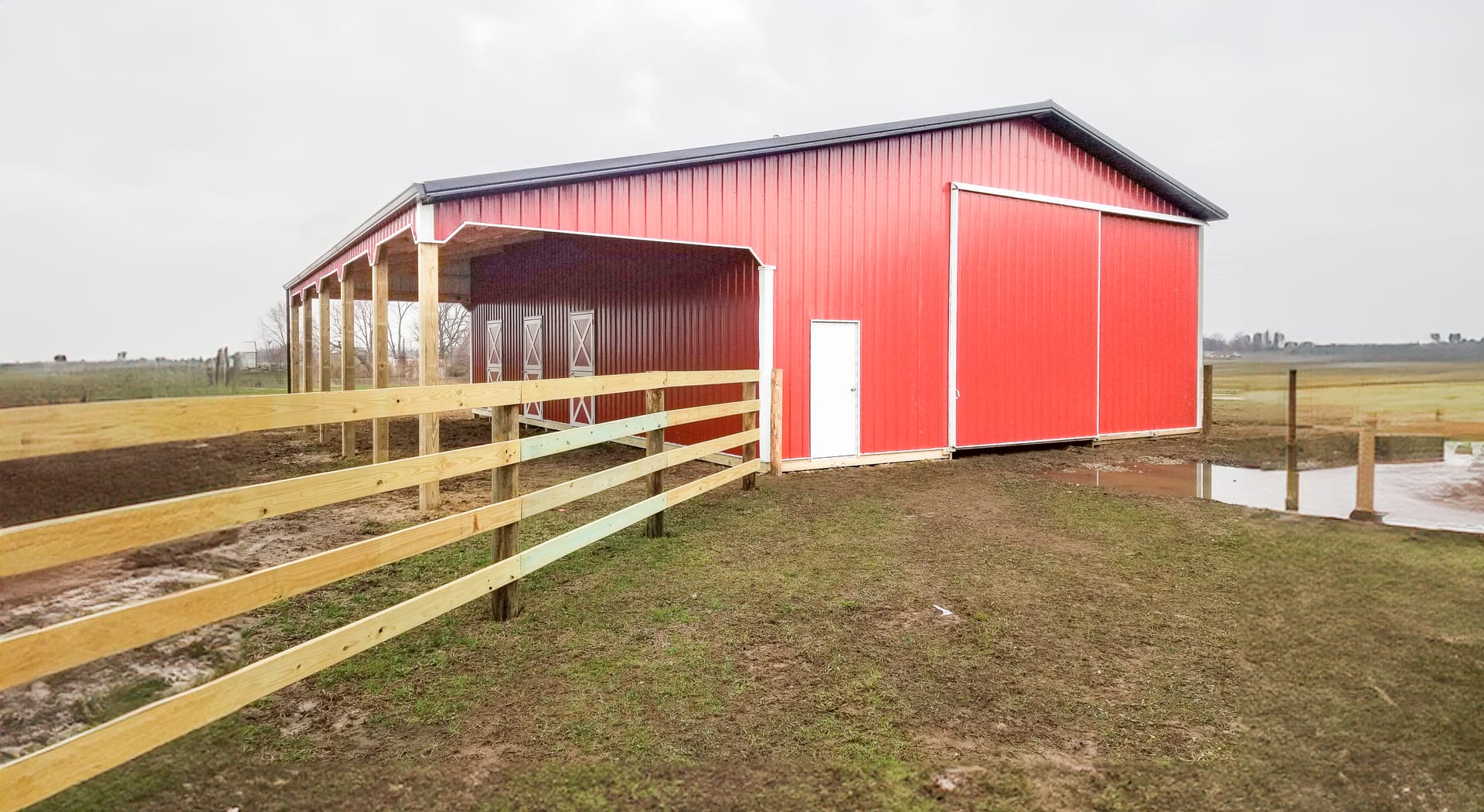 The image shows a post-frame construction building by Built-Mor Buildings, featuring a red exterior with a white door and large sliding barn-style doors. The building has a gable roof and an open covered area on one side, supported by wooden posts. It is