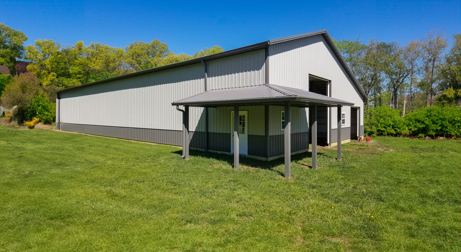 The image shows a large post-frame construction building by Built-Mor Buildings, featuring a dual-tone exterior with light gray upper walls and darker gray lower sections. A prominent feature is the covered porch with a sloped roof supported by wooden pos