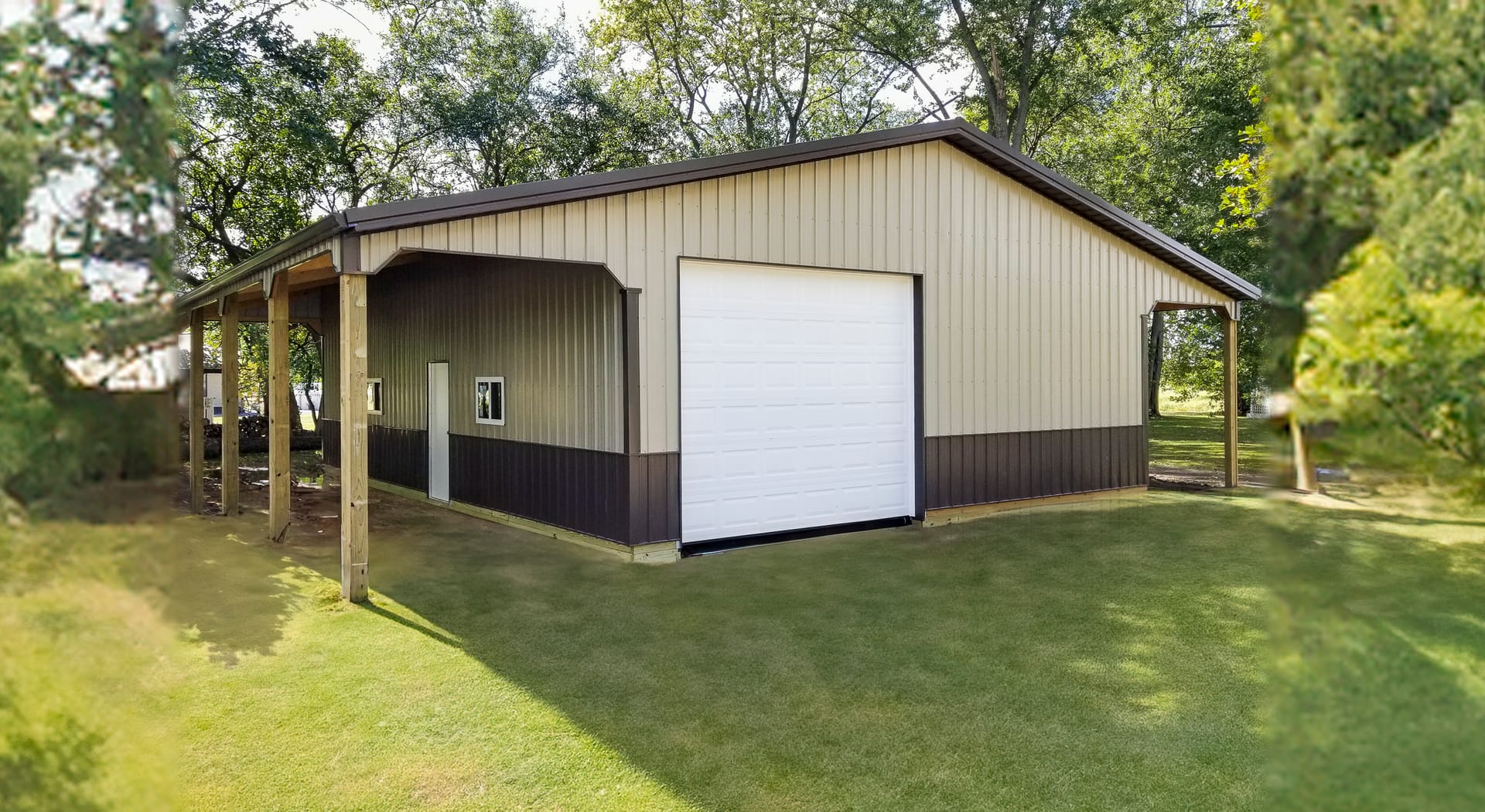A post-frame building with beige and dark brown metal siding. It features a large white overhead door, indicating potential use as a vehicle or equipment storage facility. The structure has a gabled roof and a covered open-sided extension supported by woo