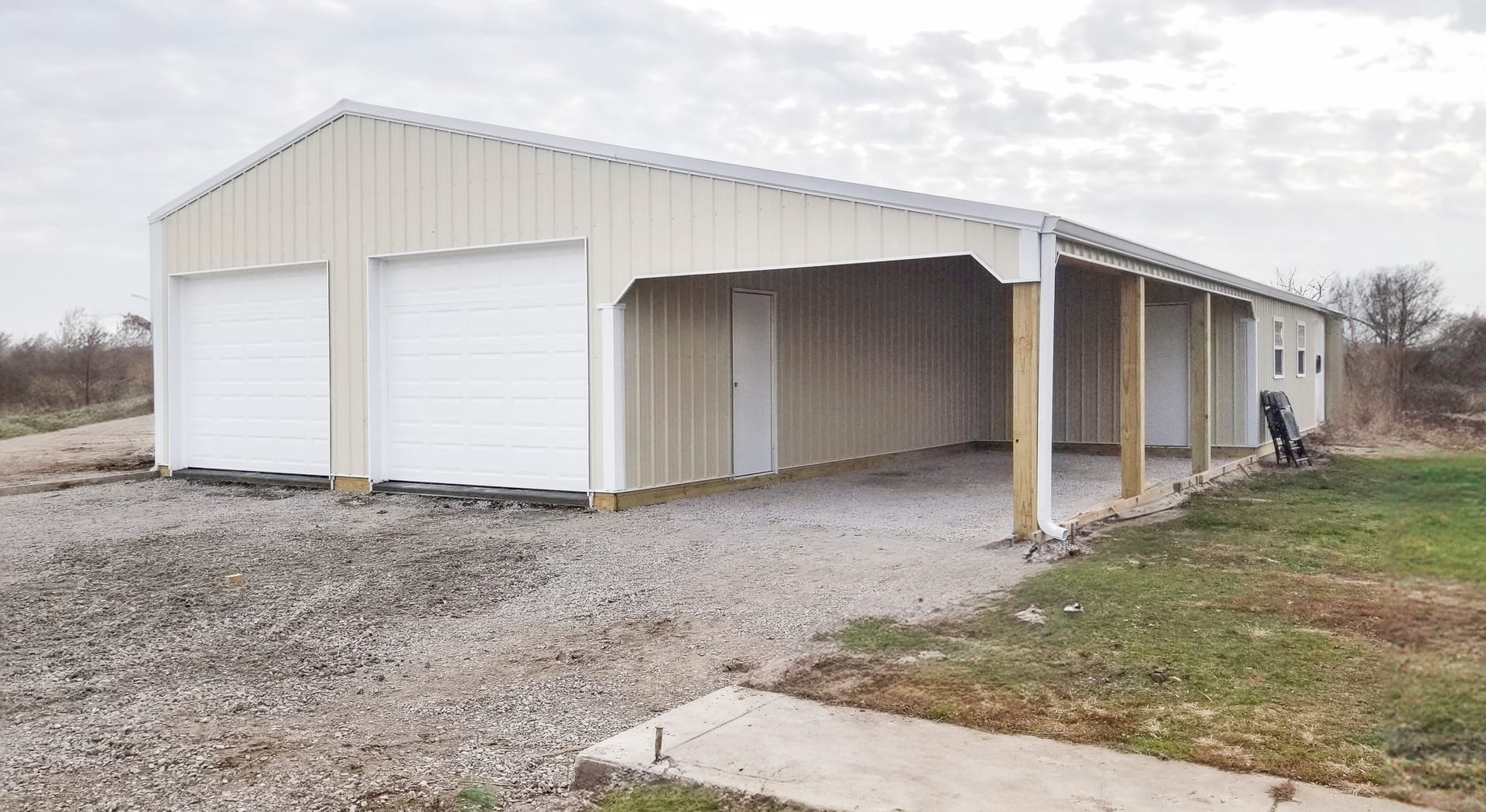 The image features a post-frame construction building by Built-Mor Buildings. It has a rectangular shape with cream-colored metal siding and a gable roof. The structure includes two large white garage doors on the left, likely for vehicle storage, and an