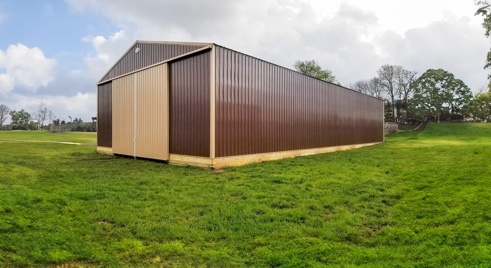 A large post-frame building with a gable roof, featuring brown metal siding and beige sliding doors at the front. Designed for agricultural or storage use, the structure sits on a grassy field. Its elongated shape and lack of visible windows emphasize fun