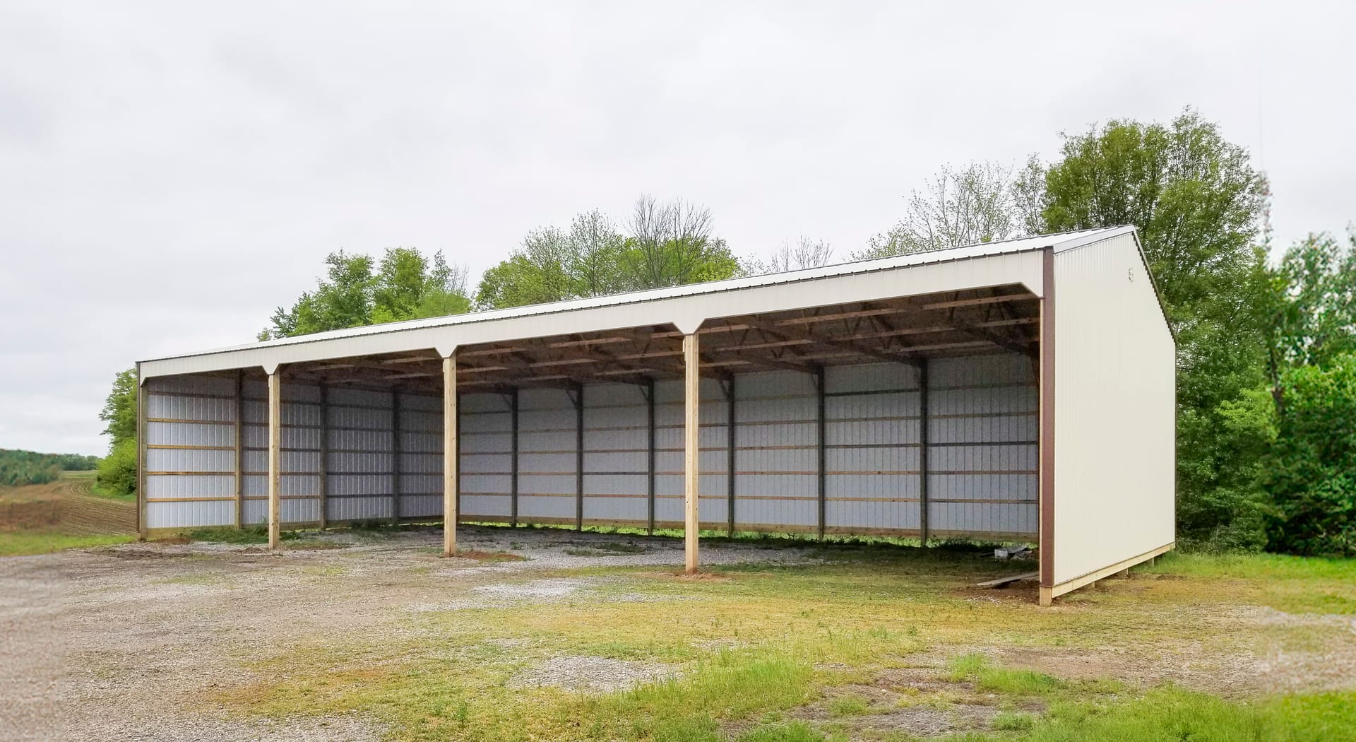 The image depicts a post-frame construction by Built-Mor Buildings, featuring a large open-sided structure likely used for equipment or vehicle storage. The building has a metal roof supported by wooden posts, with three open bays, each divided by vertica