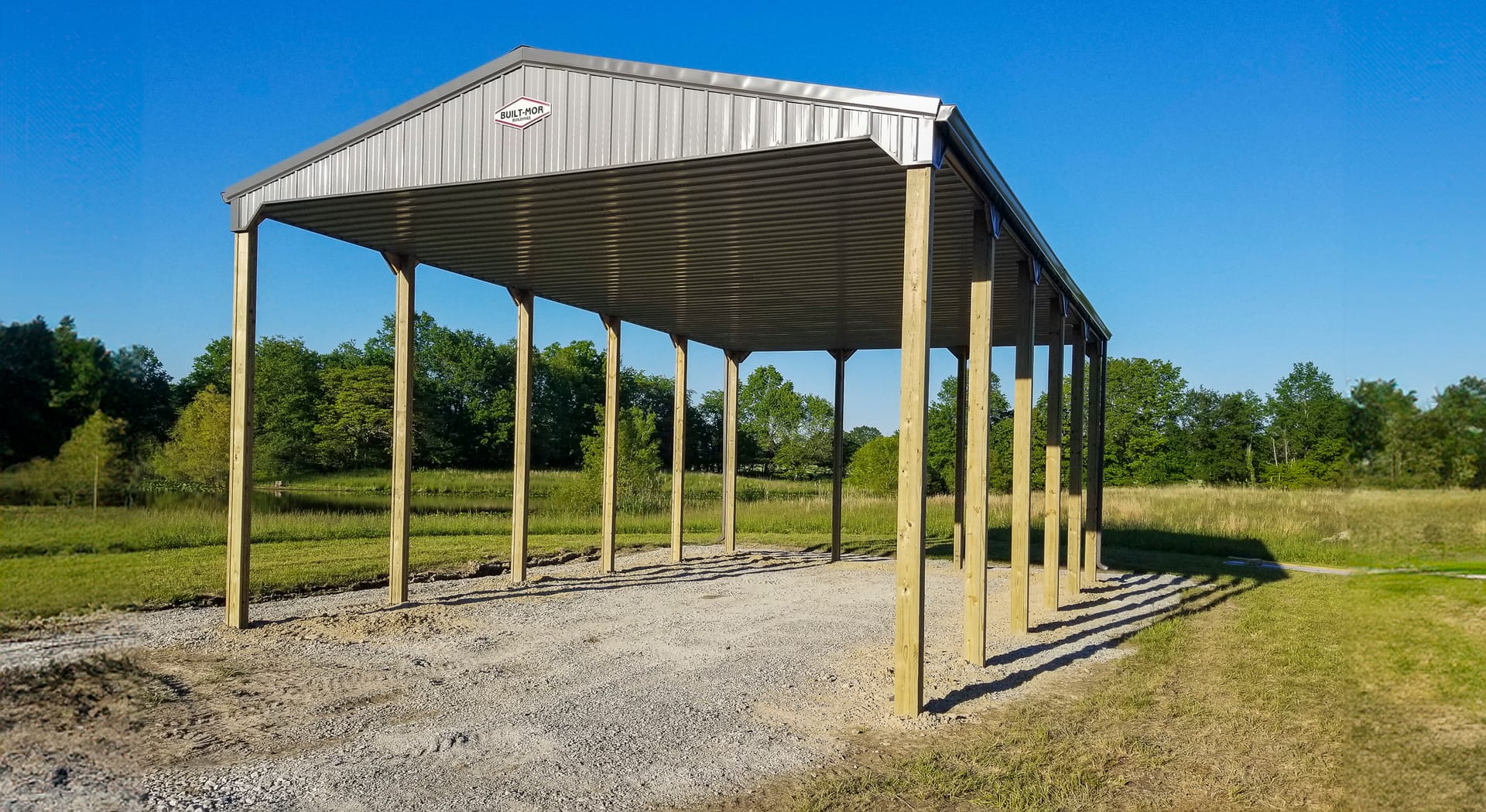 The image depicts a post-frame construction from Built-Mor Buildings, featuring a large open-sided shelter. The structure has a gabled metal roof supported by multiple evenly spaced wooden posts. It is situated on a gravel base within a grassy field. The