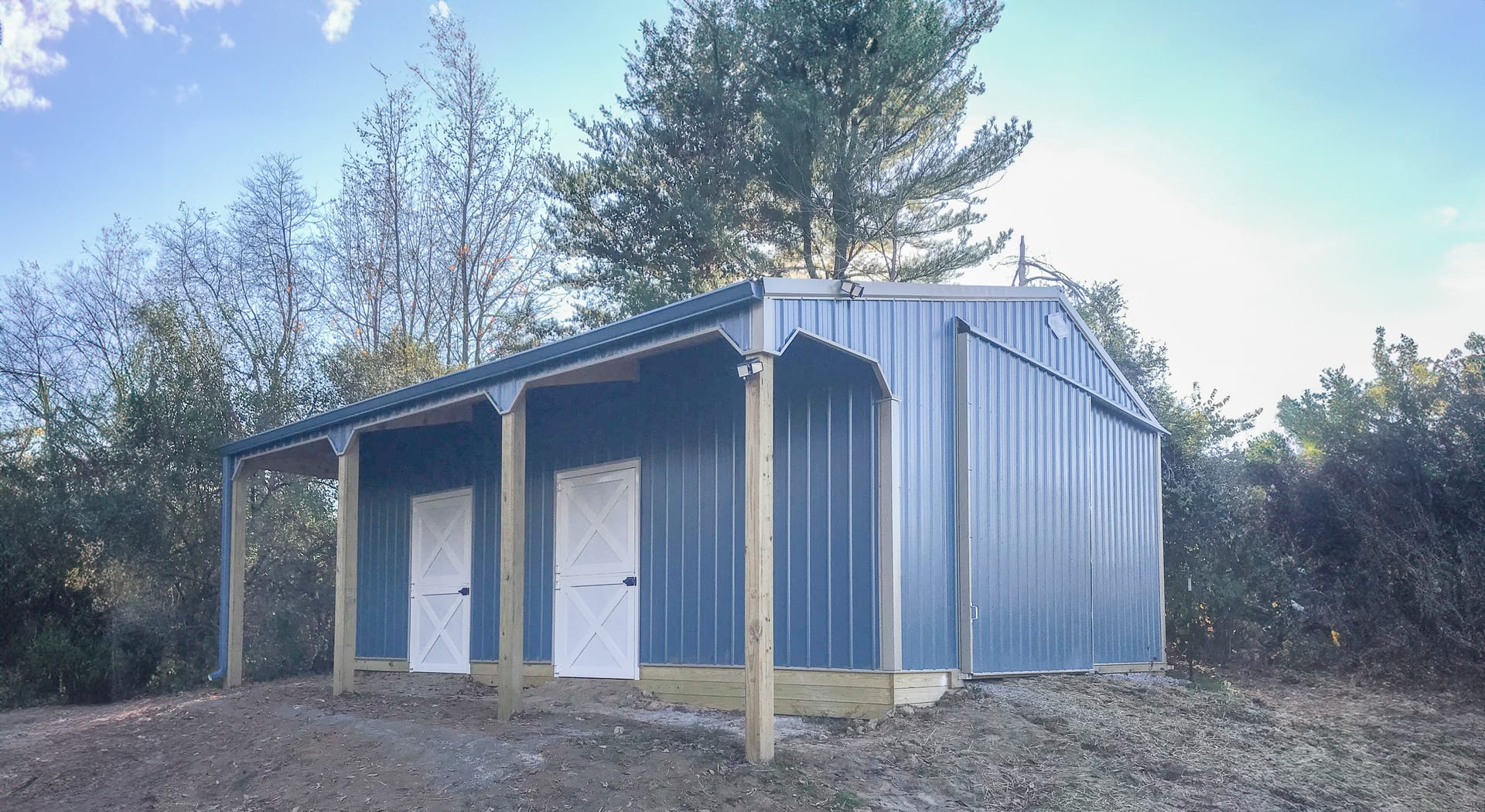 The image depicts a post-frame construction building set in a wooded area. The structure features blue metal siding and a sloped roof with a small overhang supported by wooden posts. It has two white doors on the front, indicating possible storage or util